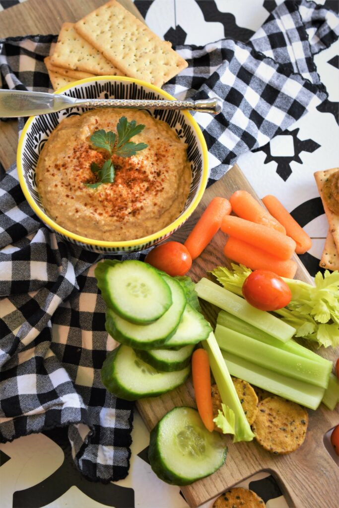 smoky chipotle almond dip in black and white yellow rimmed dip bowl on a black and white gingham napkin on wood cutting board with veggies and crackers with stainless dip spreader resting on top of bowl