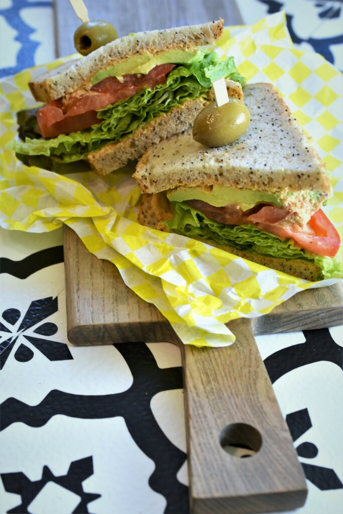 a slice veggie sandwich on the diagonal stacked off to the side garnished with an olive pick on yellow and white checkerboard deli paper on cutting board