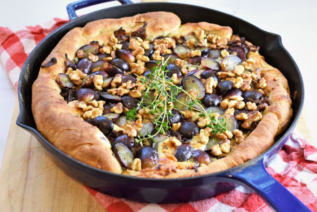 frying pan pizza with gorgonzola and walnuts with fresh thyme garnish in blue cast iron on red and white checkered napkin on wood cutting board 