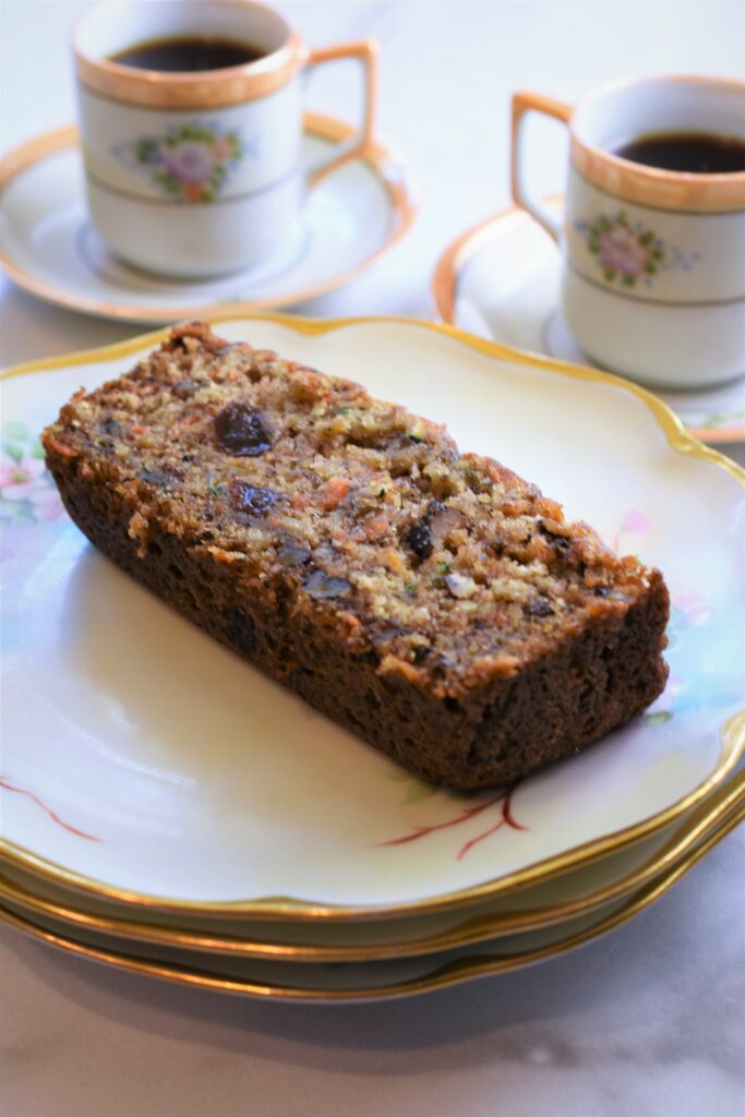 two cups of espresso in antique cups and saucers in the background with a slice of a baked treat on top of a stack of three antique floral off white and gold rimmed plates 