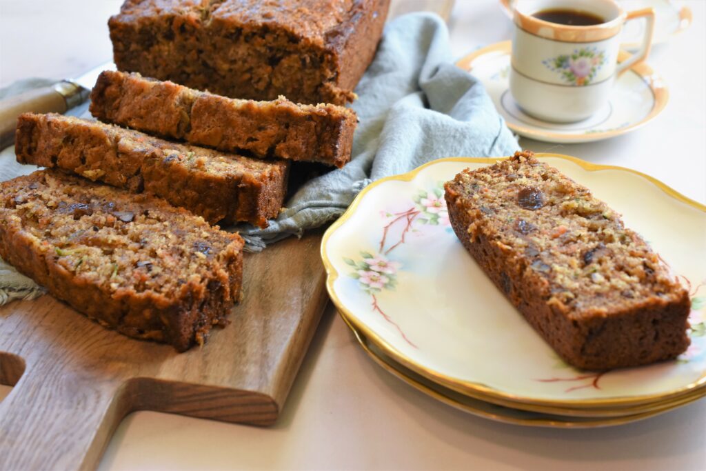 home baked vegan bread on cutting board with tattered blue cloth along side a stack of antique plates with one slice on it with a cup of espresso in the background