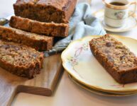 cutting board with banana zucchini carrot bread with some sliced along side antique floral plate with one slice on top and tea cup with saucer with tea in it