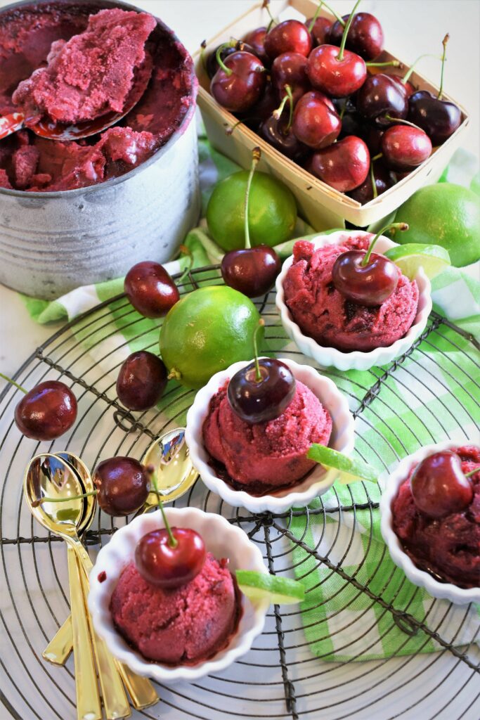 hibiscus cherry and lime sorbet in white dessert cups on wire rack with mini gold spoons on top of green and white checkered napkin with antique galvanized ice cream tin and wood basket full of cherries in the background