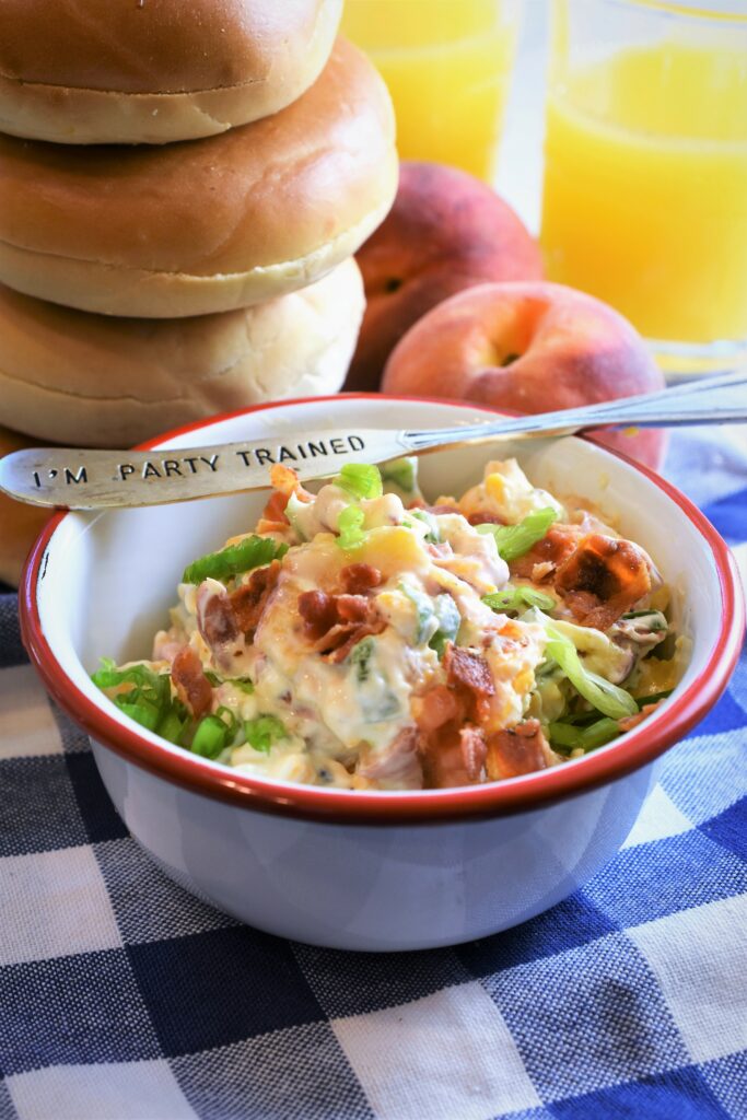 white and red enameled bowl fill with  peach bacon & jalapeno schmear next to a stack of bagels with two peaches and two glasses of orange juice in the background