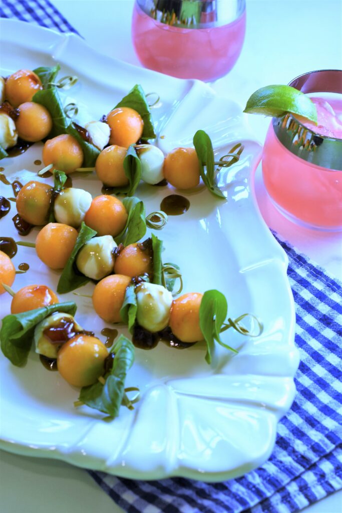 caprese salad skewer appetizer on white serving dish with cocktails with lime on the side with a blue and white gingham napkin