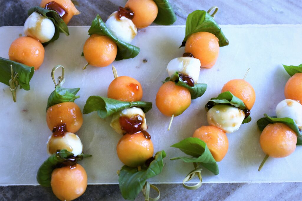 gray and white marble striped board with 7 mini skewers of melon and cheese with fresh basil displayed 
