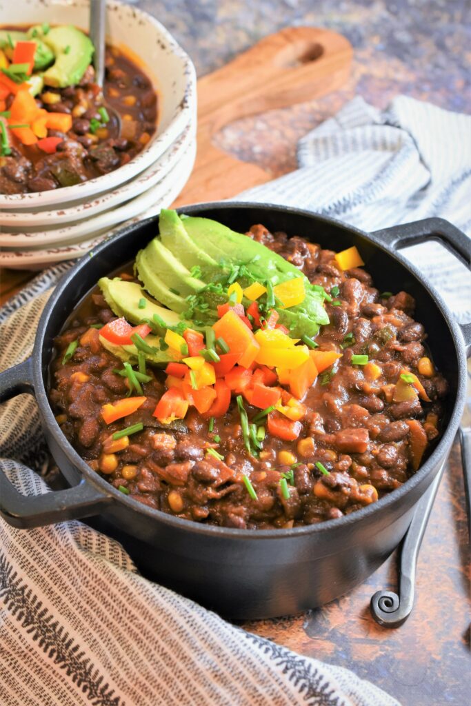 large black cast iron pot of chipotle black bean chili with coffee & cocoa and several rustic white bowls stacked in the background with the top bowl filled
