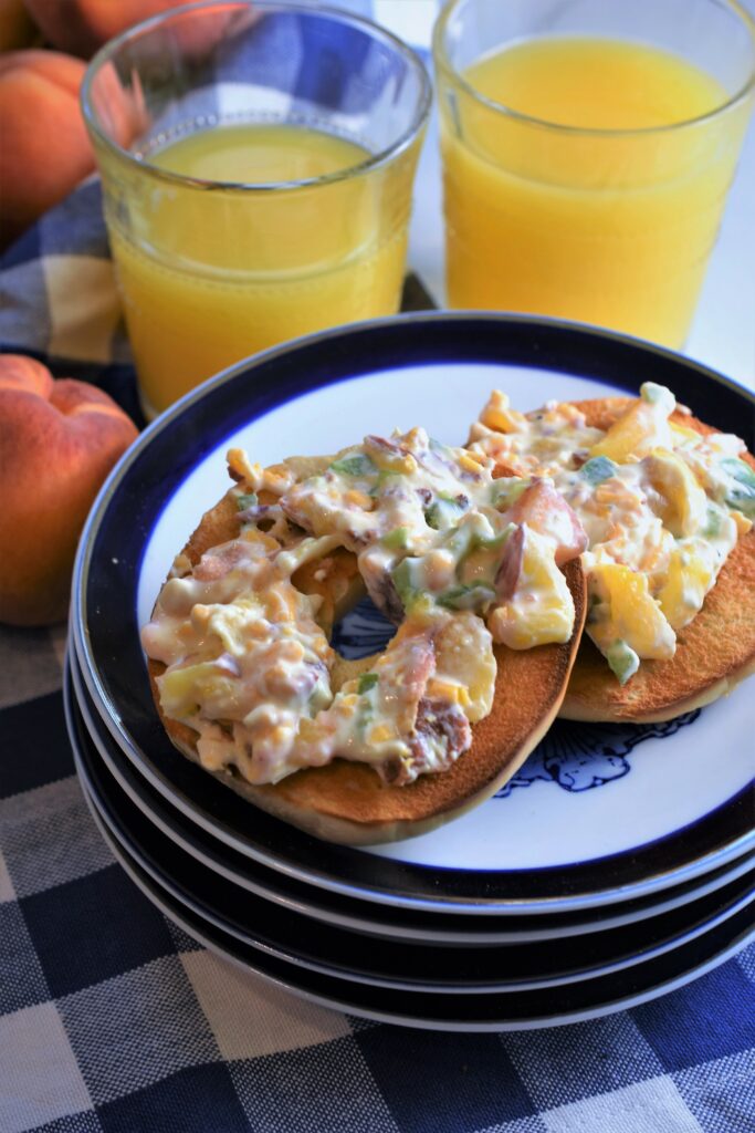 sliced and toasted bagel displayed open face schmeared with peach bacon jalapeno cream cheese with two glasses of orange juice and whole fresh peaches off to the side