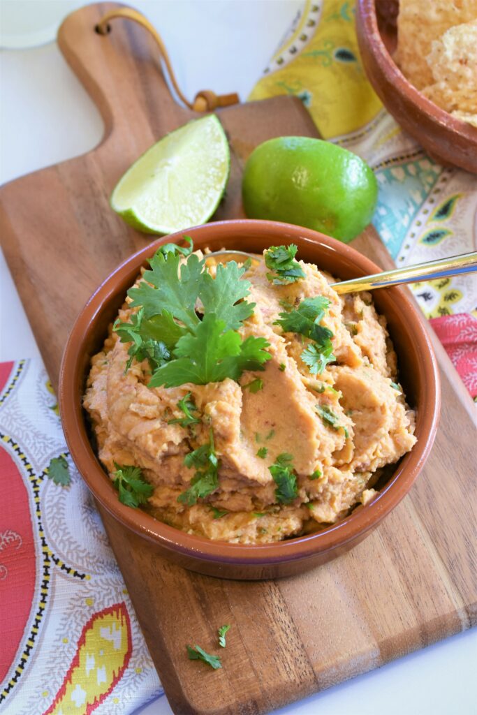 clay bowl of pureed beans with gold serving spoon with lime wedges off to the side on cutting board