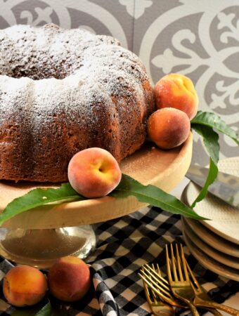 vegan banana peach Bundt cake on round wood cake stand surrounded by fresh peaches with a stack of dessert plates near by on a black and white gingham napkin in front of gray and white Spanish tile back-splash