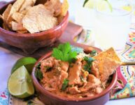 clay bowl of creamy chipotle white bean dip with tortilla chips in another clay bowl off to the side on a small wood cutting board