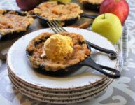 up close image of skillet snickerdoodle apple crisp on a stack of four white rustic plates with iron handled fork