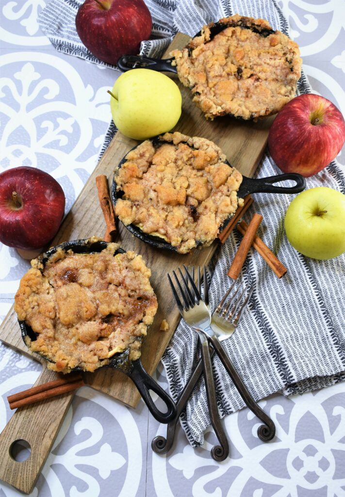 rectangle cutting board with small iron skillets of snickerdoodle apple crisp with black iron handled forks, apples and cinnamon sticks in the background