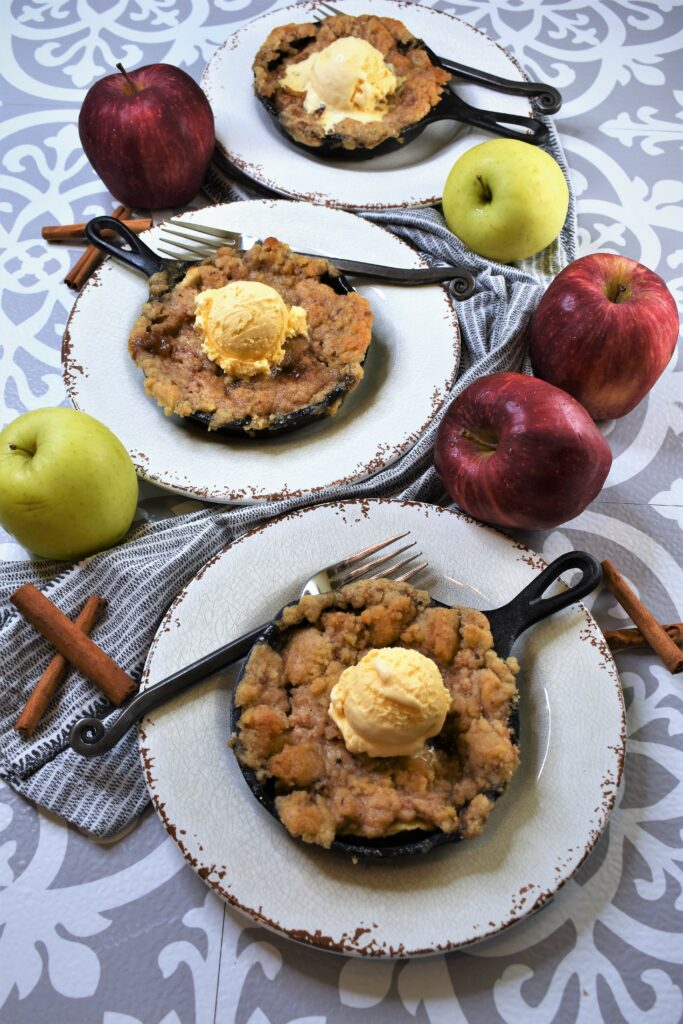 overhead shot of several small cast iron pans with apple crisp along with apples and cinnamon sticks in the background on gray and white spanish tile countertop
