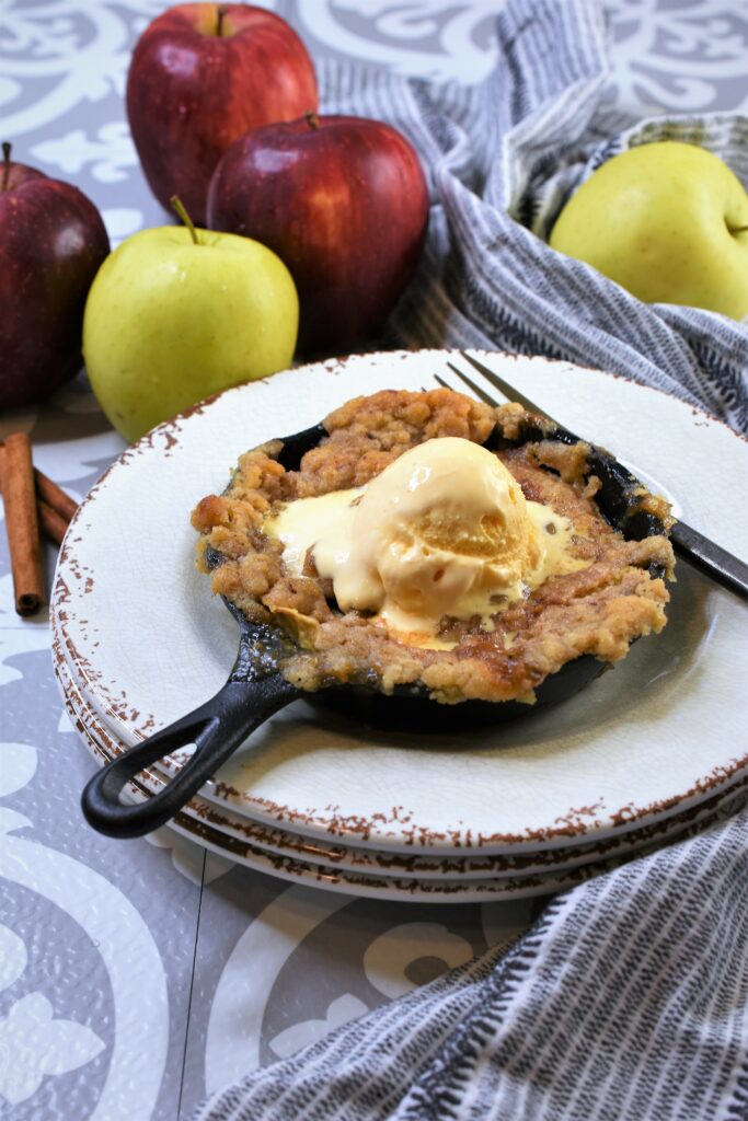 snickerdoodle apple crisp baked in individual cast iron skillet on a stack of rustic white plates with a scoop of ice cream on top with red and yellow apples in the background