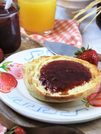 red pepper strawberry jam on english muffins on strawberry printed plate on cutting board with strawberries in the background along with full open jar of jam on orange and white gingham printed napkin