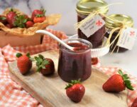 red pepper straberry jam in glass serving jar with white porcelain spoon on cutting board with many strawberries scattered around