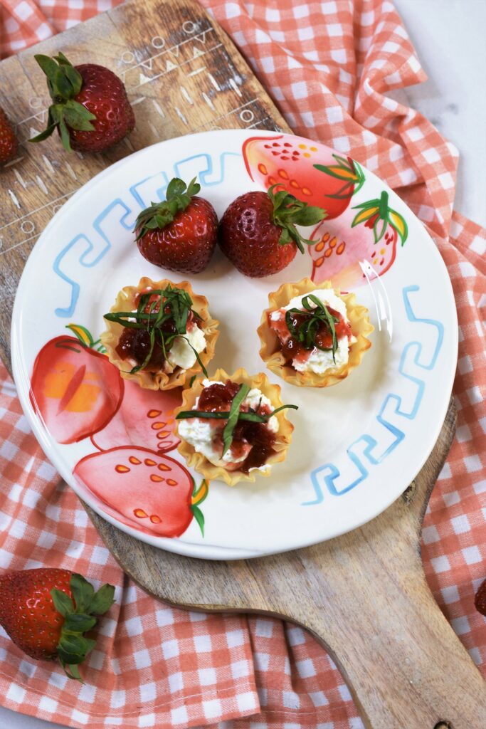 a strawberry printed ceramic plate with phyllo cups filled with cream cheese with red pepper strawberry jam drizzled on top of each one with chopped basil as garnish on top of cutting board