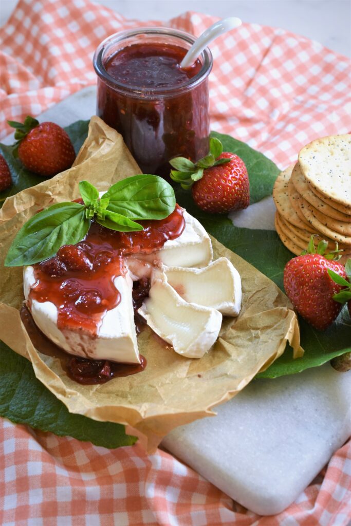 a cut wheel of Brie on parchment paper on marble cutting board with jam poured on top along side of a stack of crackers with strawberries and an open jar of jam