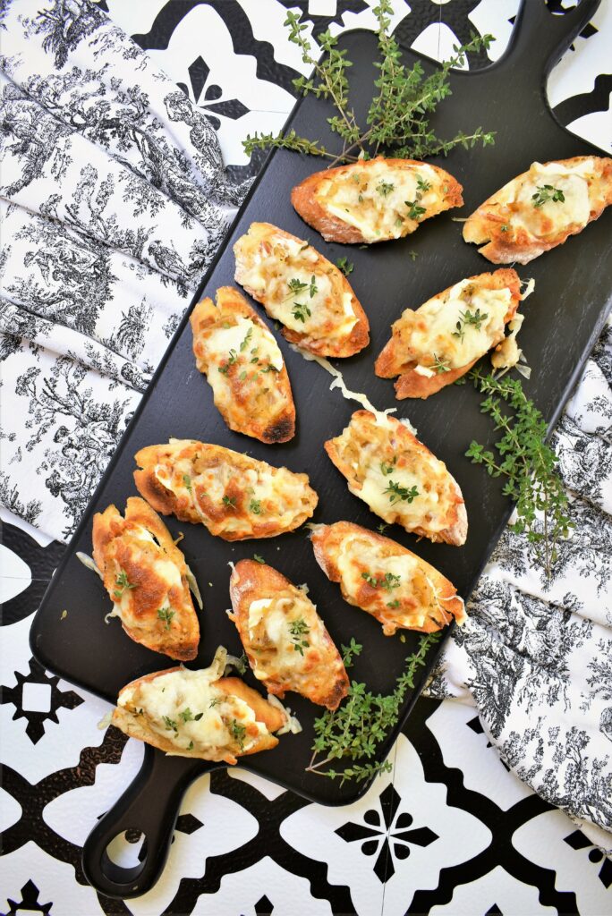 brandied caramelized onion swiss crostini on black cutting board on black and white tile countertop with French toile fabric