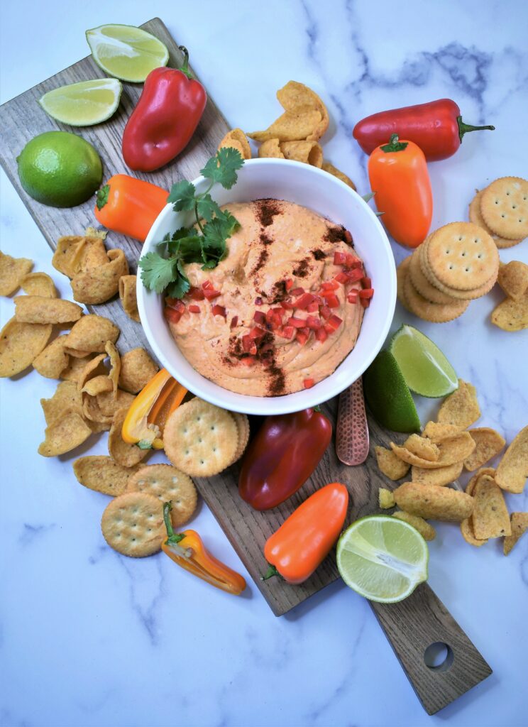 chipotle lime cheddar dip in white bowl on cutting board with crackers, corn chips and mini peppers scattered around